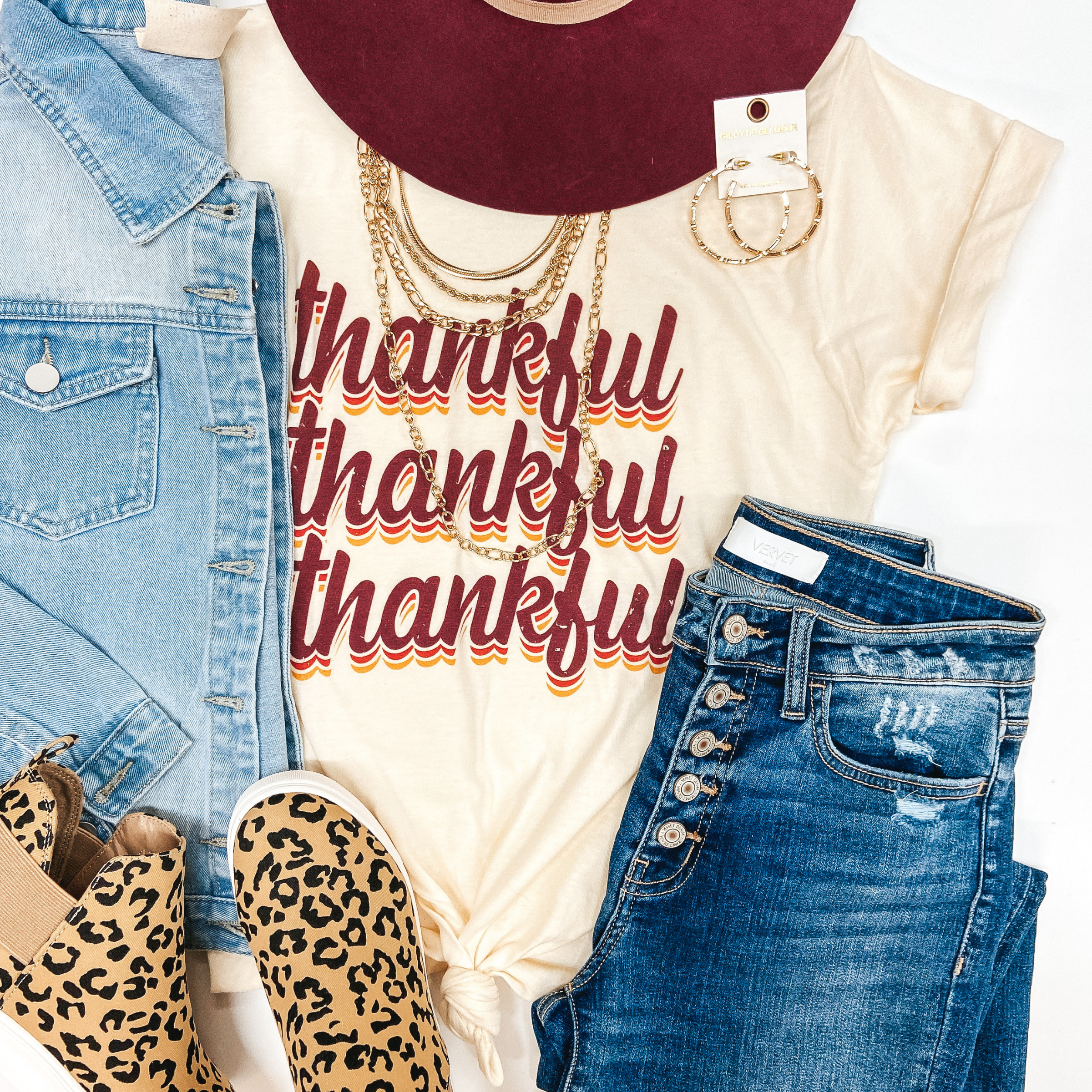 Ivory white tee with the words "thankful" written three times stacked in a burgundy color and orange then yellow shadow. Other clothes to make a full outfit are styled around the tee including a burgundy Charlie One Horse Hat, hoop earrings, necklaces, light wash denim jacket, medium wash jeans, and leopard print sneakers.