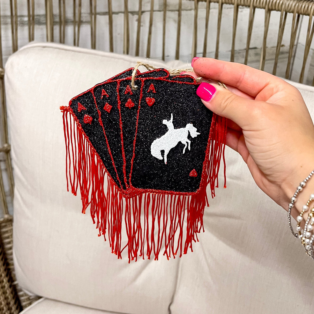 Hand holding a black and red playing card freshie with red fringe on a beige cushioned chair.