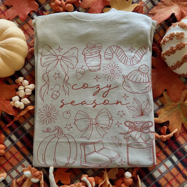 Folded tee with autumn-themed design on a bed of fall leaves and pumpkins.