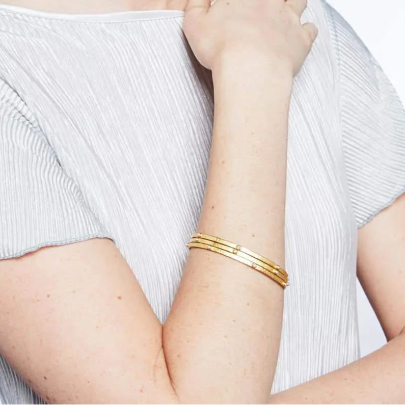 Woman wearing a white top and gold bracelets with pearls on a plain background.