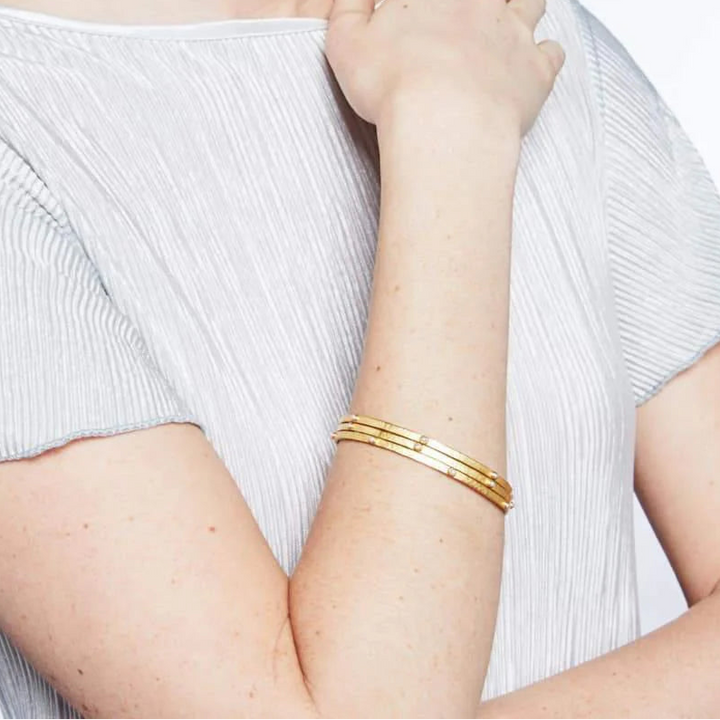 Woman wearing a white top and gold bracelets with pearls on a plain background.