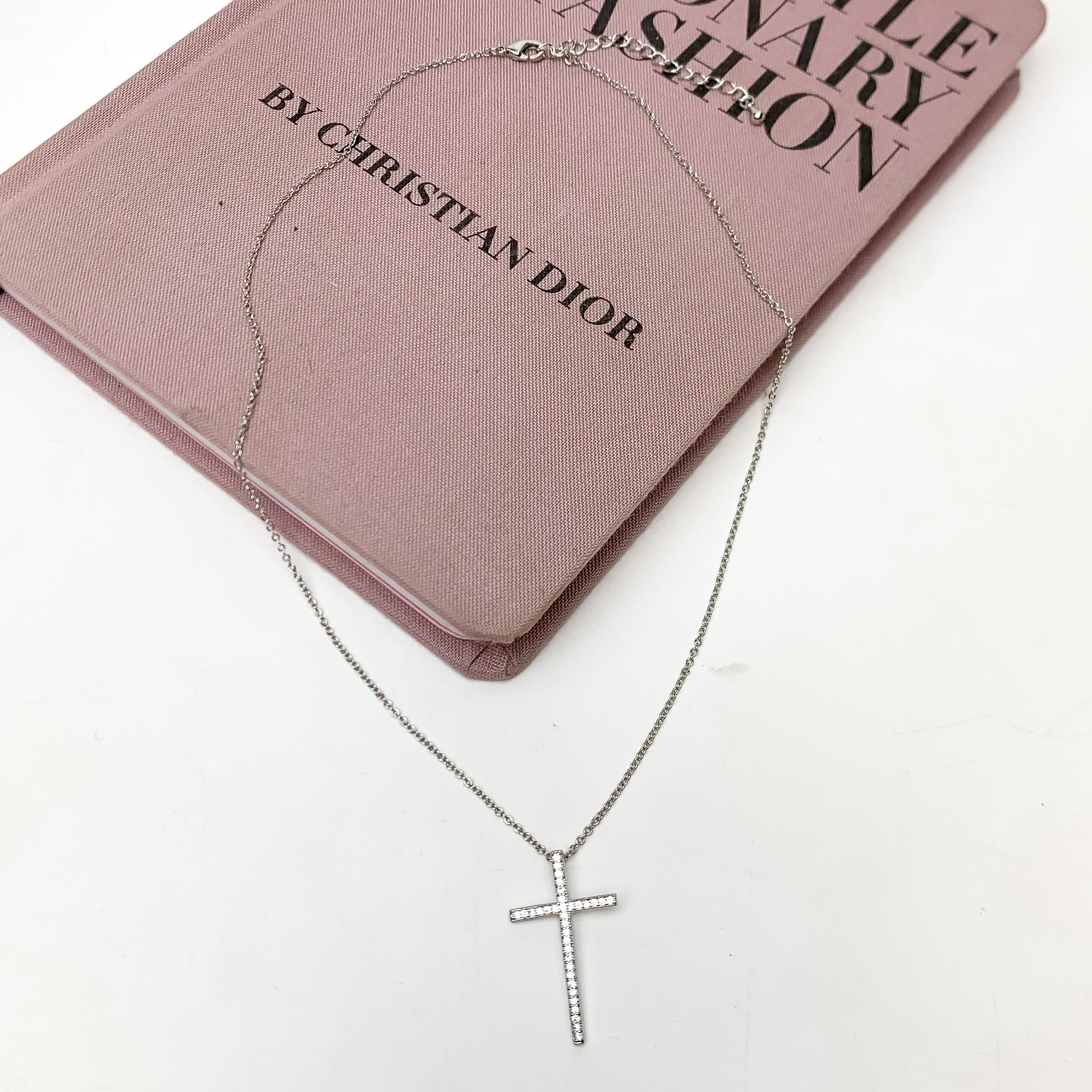 Silver Tone Chain Necklace With Clear Crystal Cross. This necklace is pictured on a white background with part of it on a pink book.