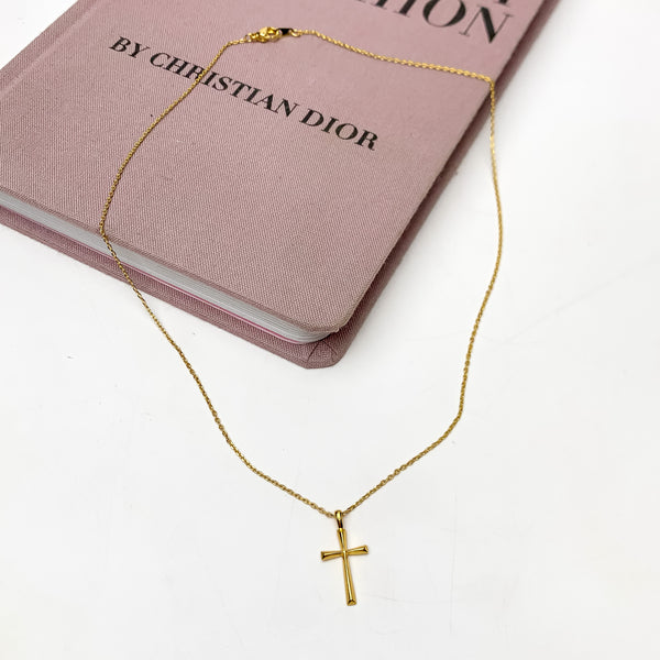 Gold Tone Glory Chain Necklace With Cross Pendant. This necklace is pictured on a white background with part of it on a pink book.
