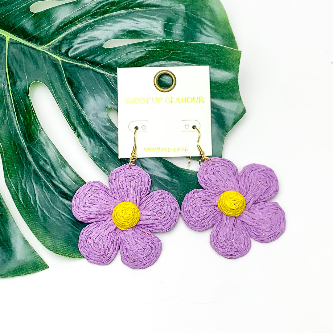 Darling Daisy Raffia Wrapped Flower Earrings in Purple. Pictured on a white background with the earrings laying on a large leaf.