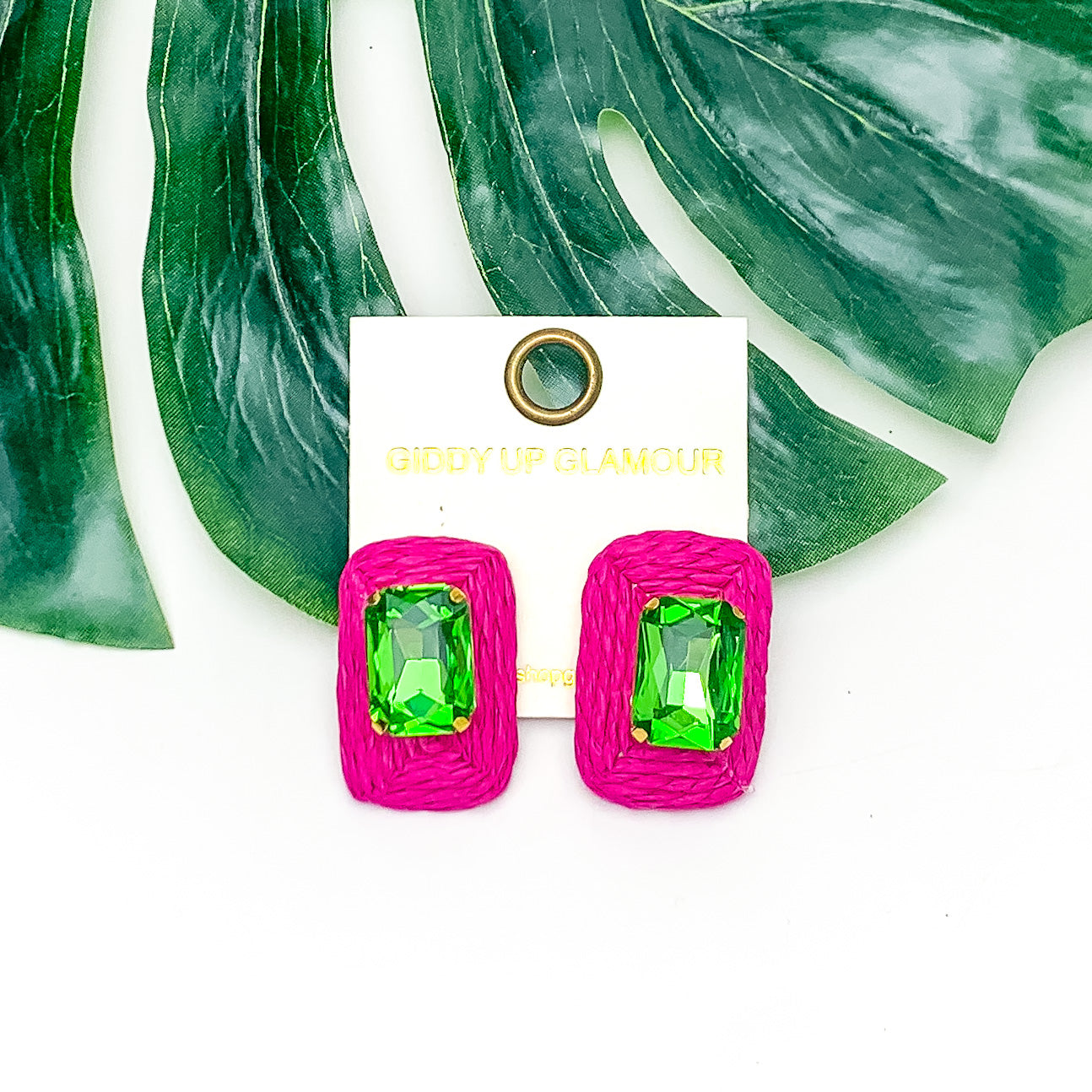 Truly Tropical Raffia Rectangle Earrings in Hot Pink With Green Crystal. Pictured on a white background with the earrings laying on a large leaf.