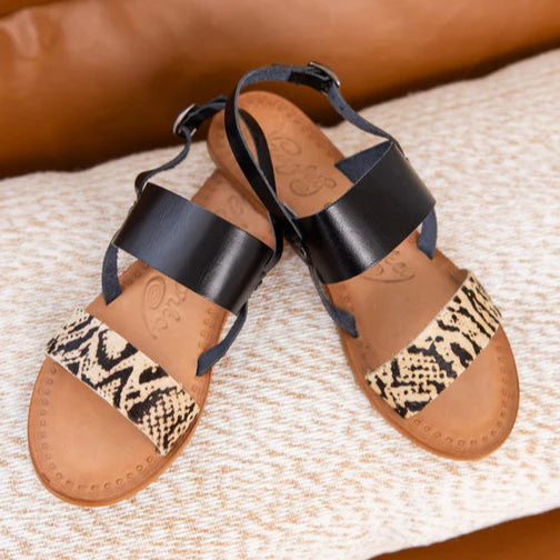 A tan and white background with strappy sandals. The sandals are on a brown sole with the toe strap in a snakeskin print and the second strap leading to the back ankle strap are solid black.
