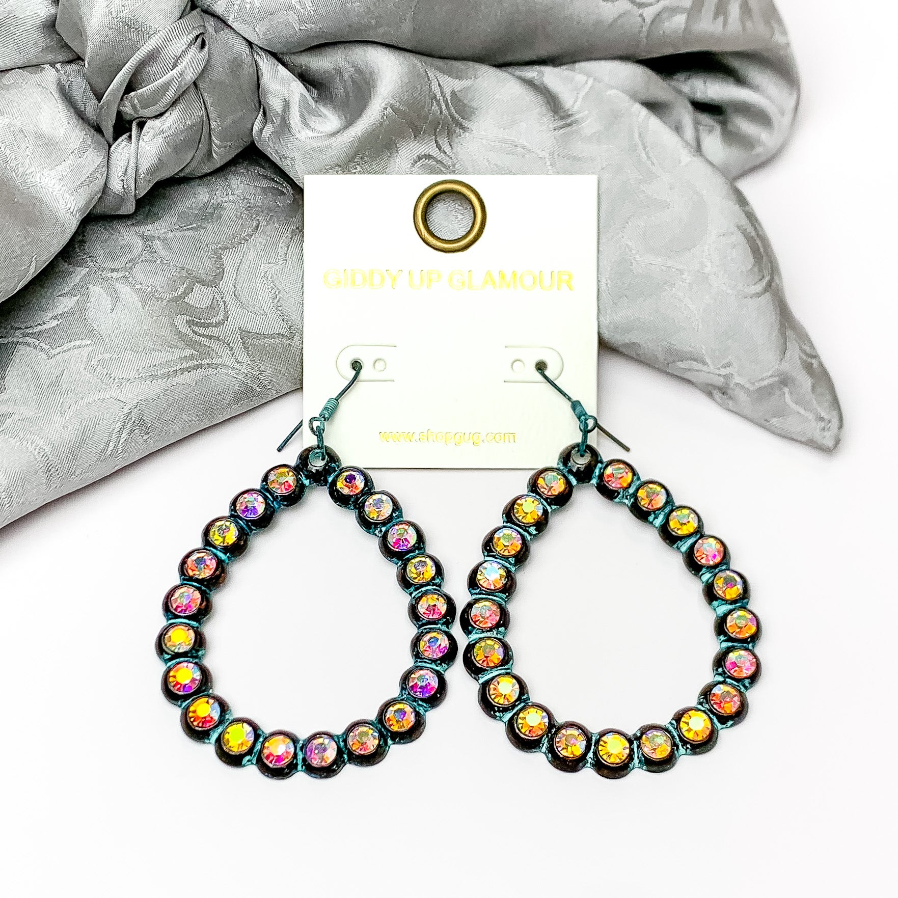 Open Teardrop Earrings with AB Crystal Outline in Patina. Pictured on a white background with sliver fabric at the top.