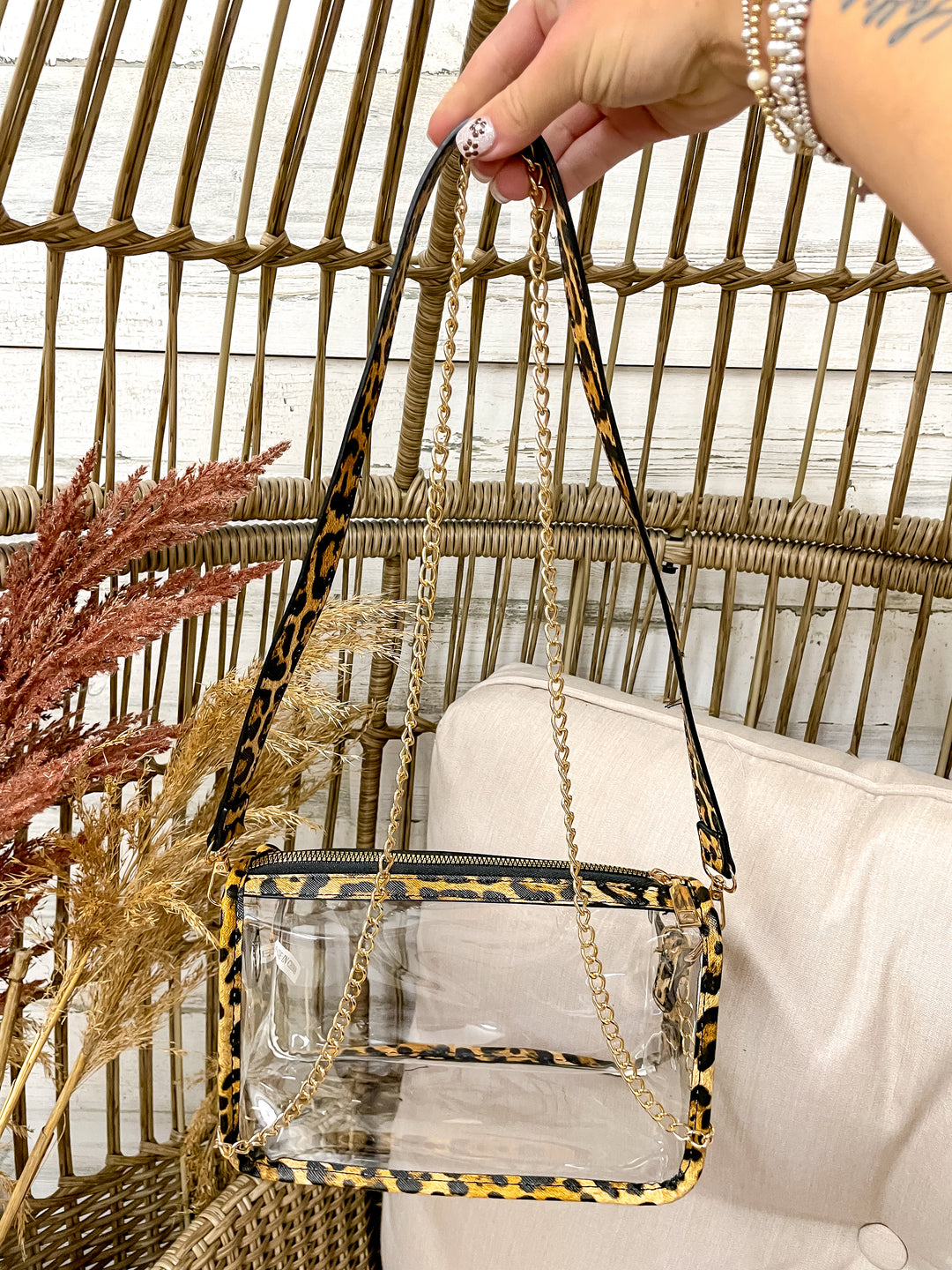 Clear handbag with leopard print strap held by a hand against a wicker chair and decorative plants background.