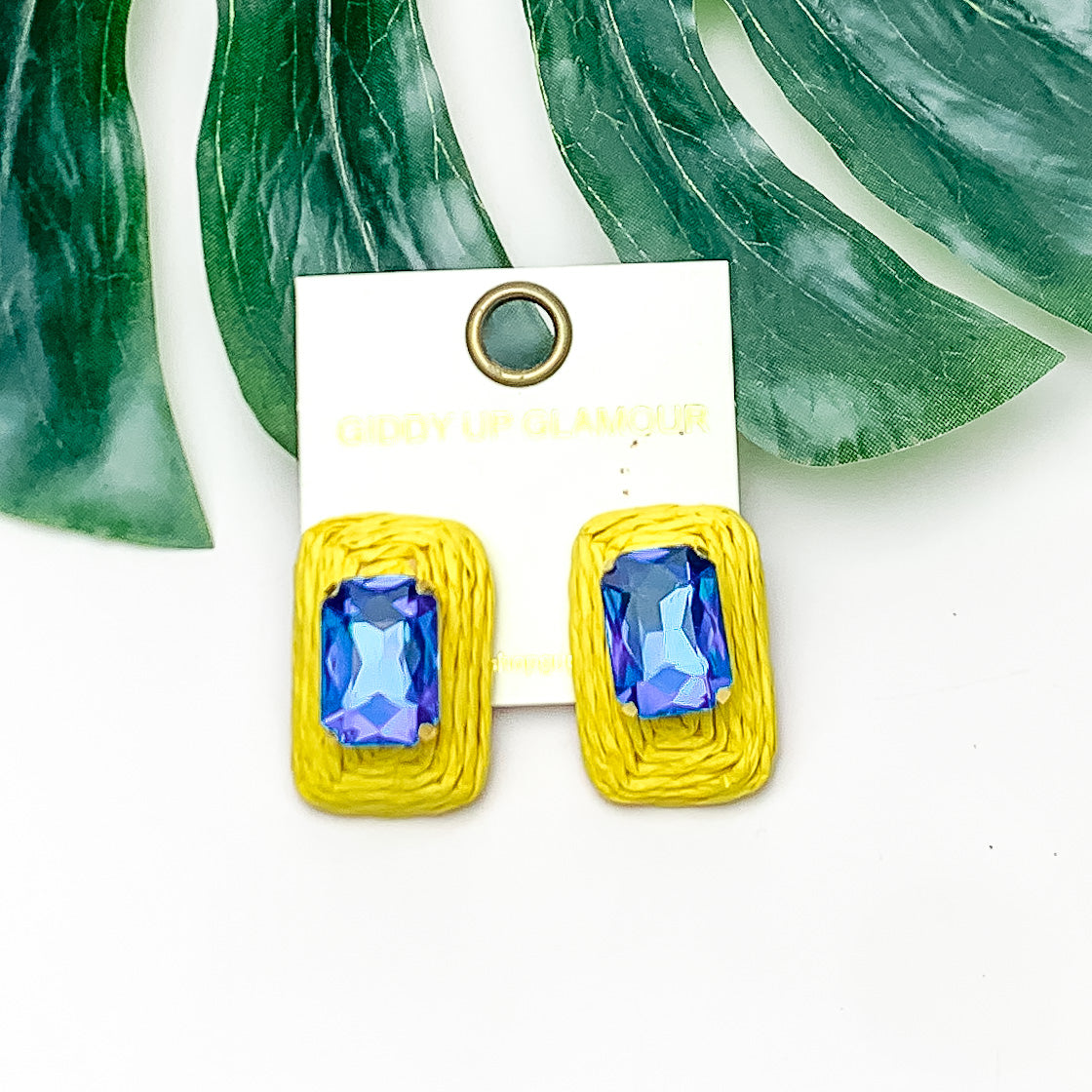 Truly Tropical Raffia Rectangle Earrings in Yellow With Blue Crystal. Pictured on a white background with the earrings laying on a large leaf.