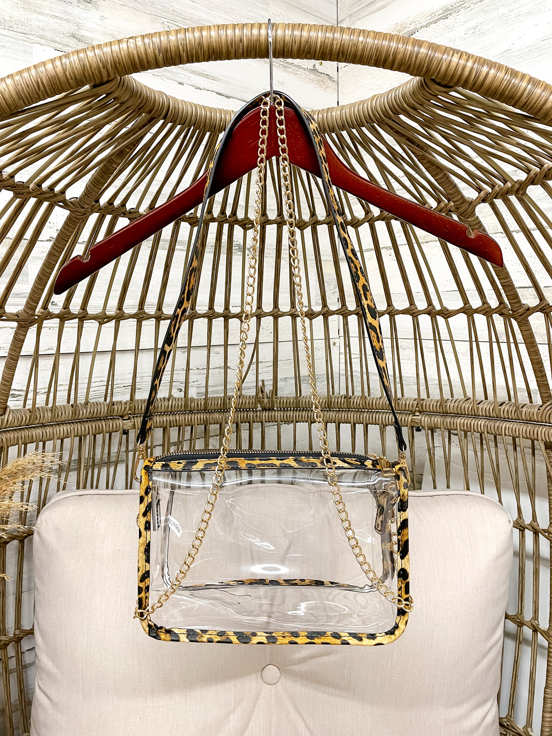 Clear handbag with leopard print accents hanging on a red hanger against a wicker chair background.