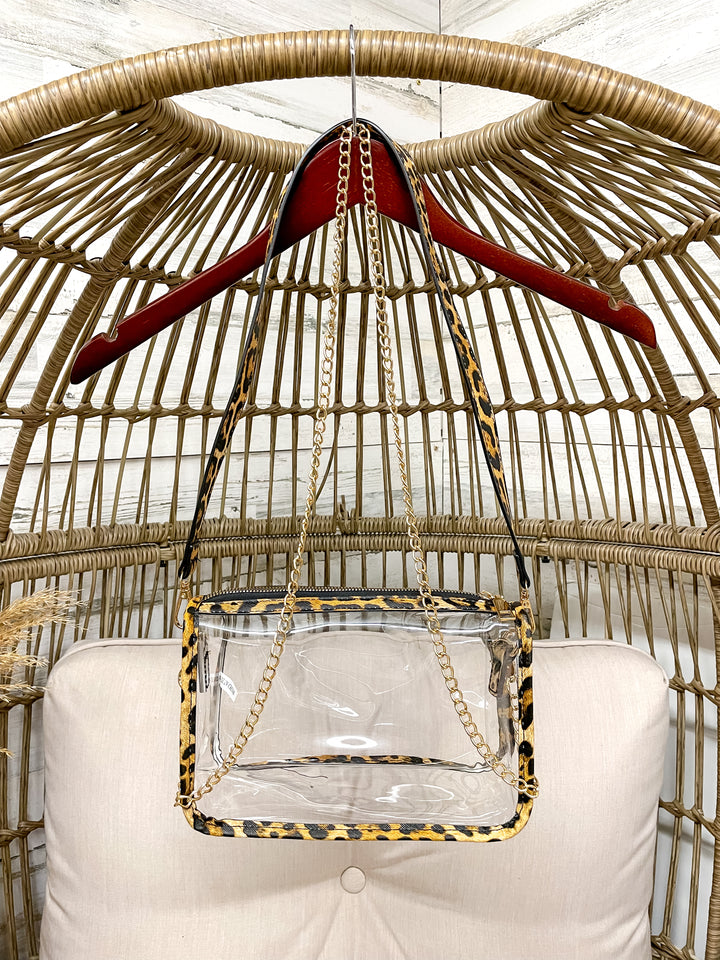 Clear handbag with leopard print accents hanging on a red hanger against a wicker chair background.