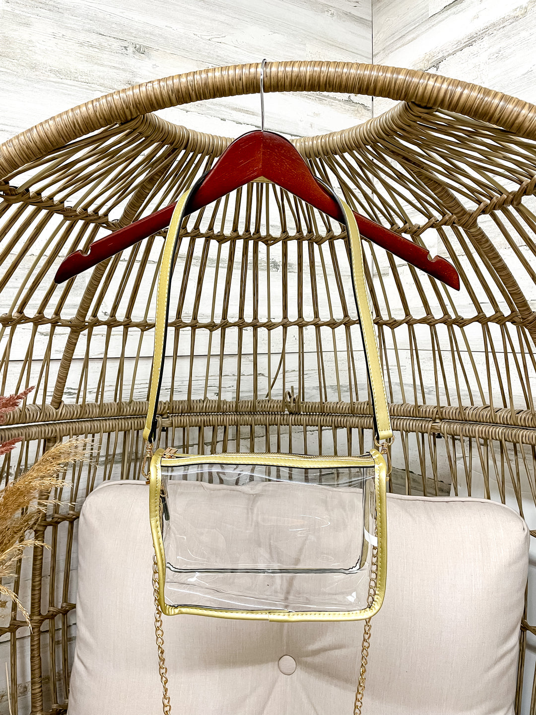 Clear handbag with gold chain strap hanging on a red hanger inside a wicker chair.