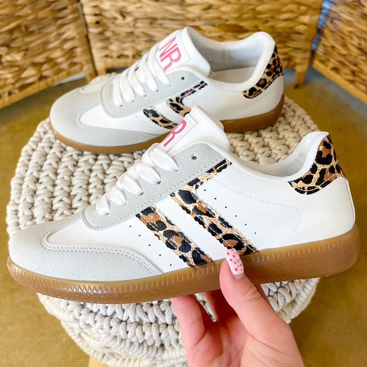 White sneakers with leopard print accents held by a hand on a woven stool.