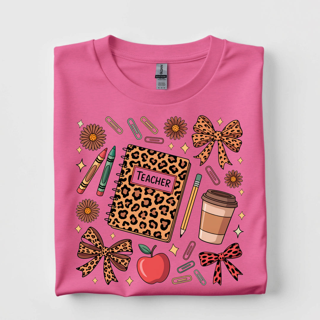 Hot pink graphic tee featuring a leopard print teacher notebook surrounded by classroom supplies, coffee, bows, flowers, crayons, and an apple.