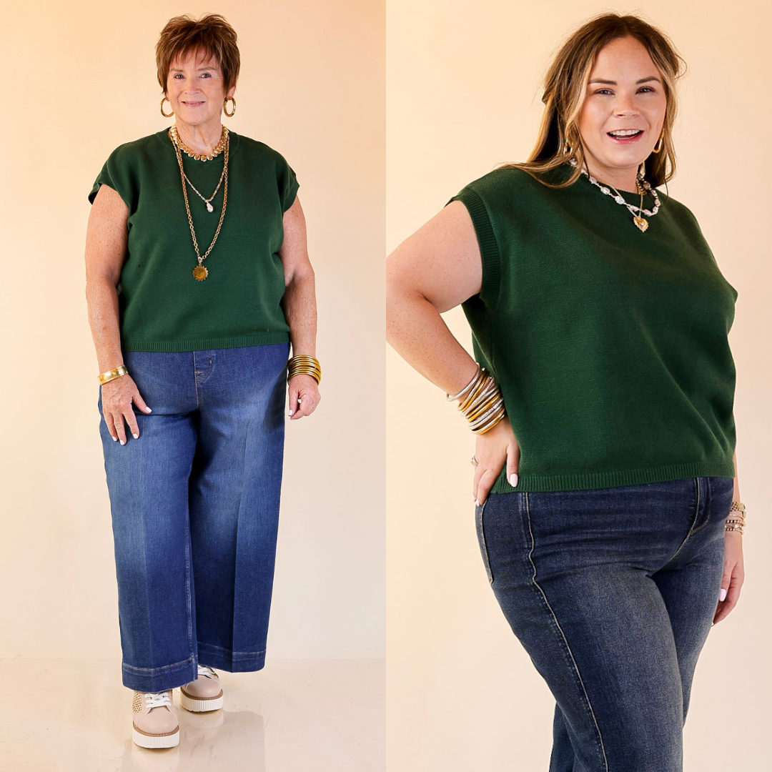 Two women wearing forest green sweater tops and blue jeans against a beige background