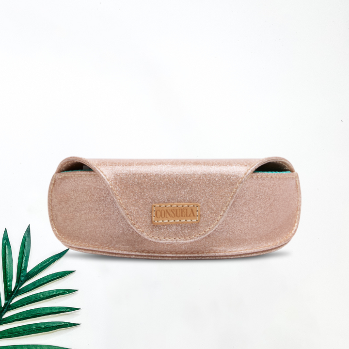 A solid white background with palm tree leaf and a sunglass case by Consuela. Emery cloth brings all the sparkle! With its gold glitter exterior, Magnetic closure, and genuine leather Consuela logo. The interior has a Soft mesh lining to protect our sunglasses!