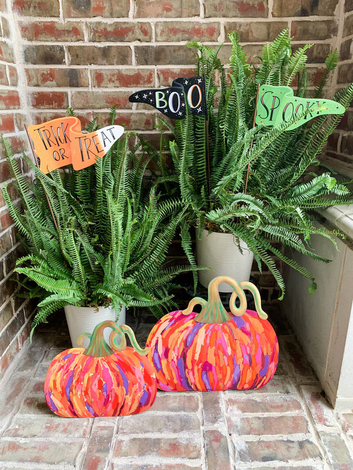 Pumpkin-shaped decorative decor pieces with colorful painted stripes in orange, pink, purple, and blue, styled with potted ferns and autumn signs on a brick floor against a brick wall.
