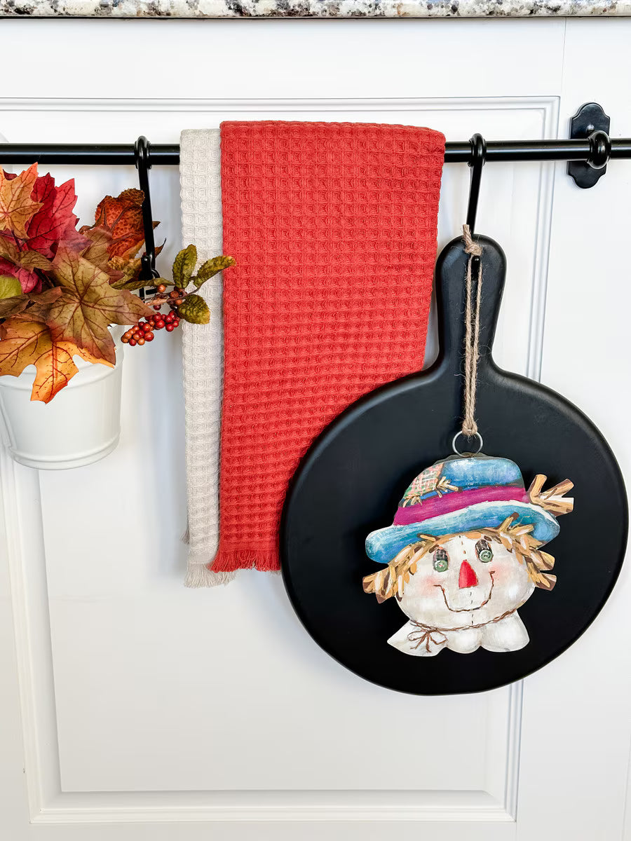 Round Top Collection Scarecrow Charm hanging on a black kitchen display with a turquoise and pink hat and colorful fall details.