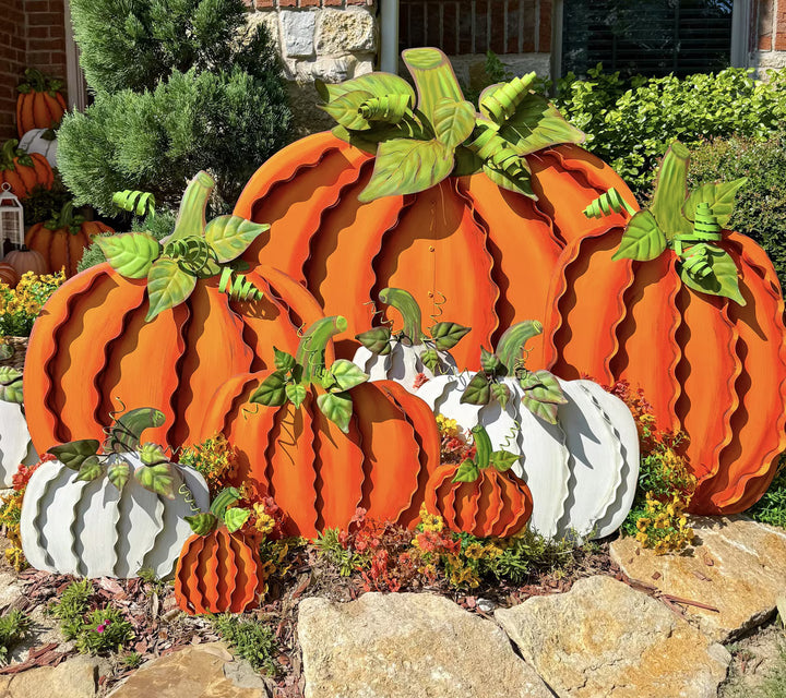 A decorative autumn yard display featuring large and small corrugated metal pumpkins in orange and white, arranged outdoors in front of a house with shrubs and stones.