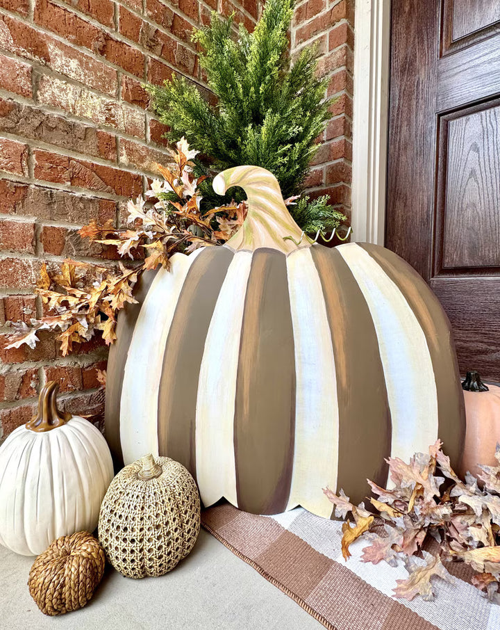 A large decorative striped pumpkin on a front porch, painted in vertical bands of cream, taupe-brown, and white, with a faux green pine top and autumn leaves around it. Smaller pumpkins and a brick wall are visible.