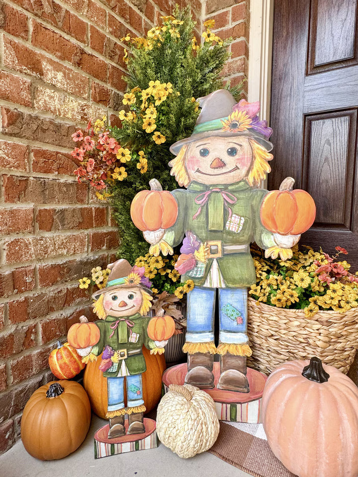 Two autumn scarecrow yard decorations displayed on a brick porch next to pumpkins and flower baskets. The larger scarecrow stands on a wooden platform holding two small pumpkins, while the smaller one stands beside it holding two even smaller pumpkins.
