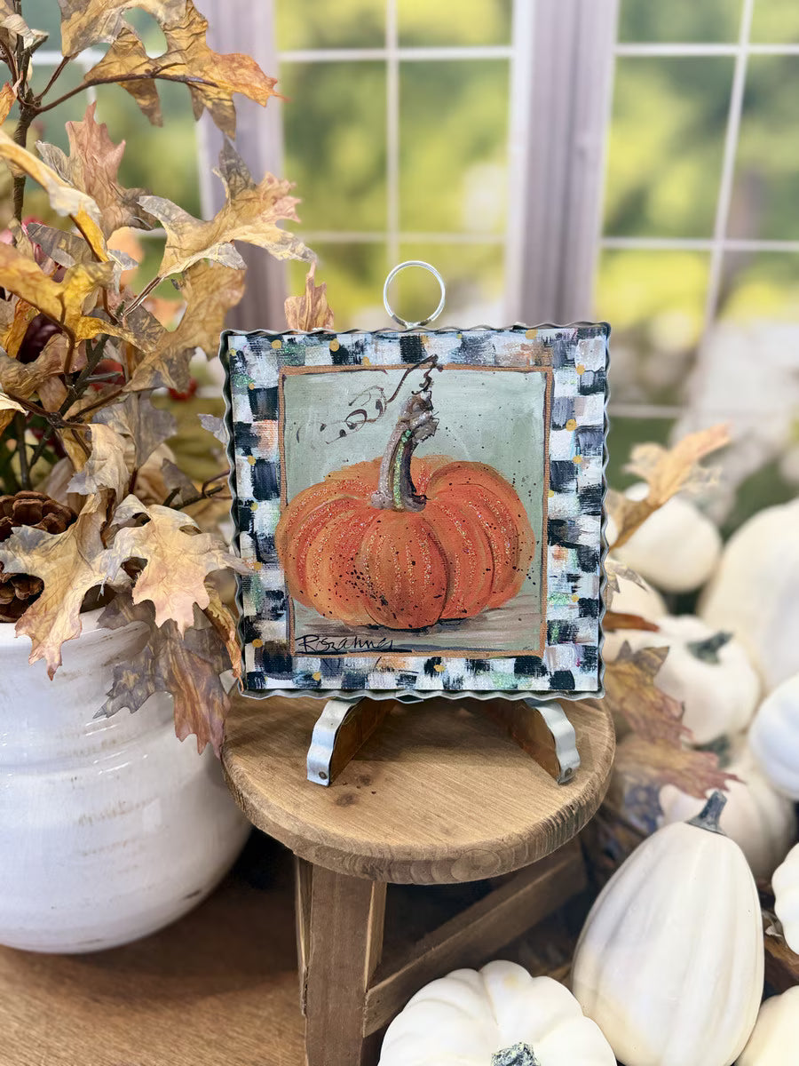 A small square decorative art piece featuring a painted orange pumpkin, displayed on a wooden stand in a fall-themed setting with pumpkins, dried leaves, and a window in the background.