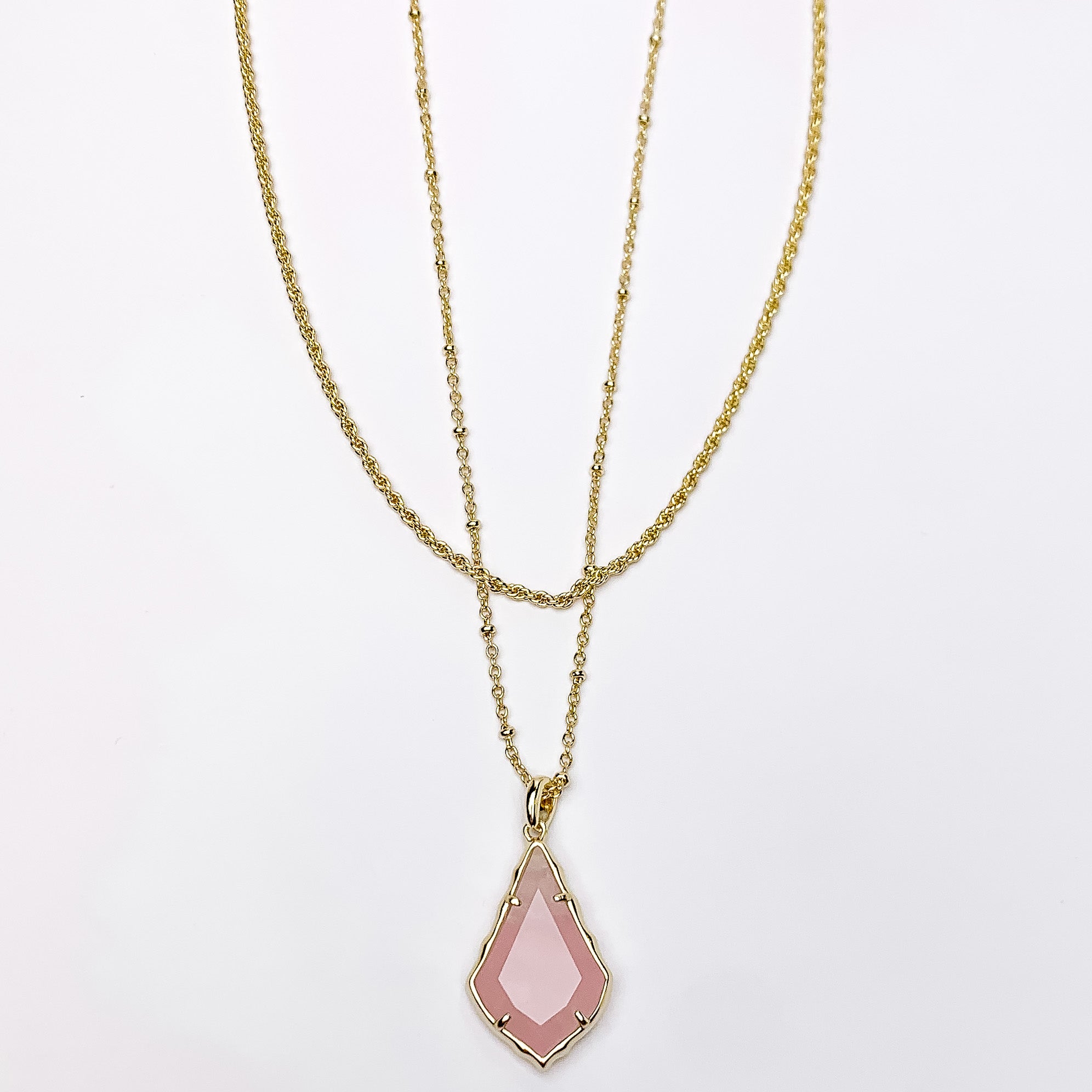 Two gold chain necklaces with the longer strand that has a gold and rose quartz pendant. This necklace is pictured on a white background. 