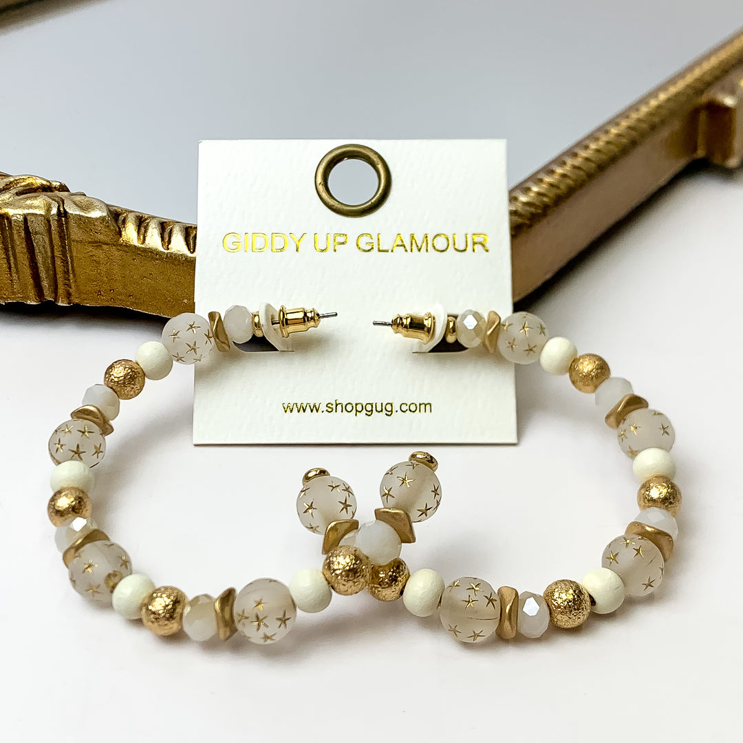 A pair of white and gold hoop earrings featuring gold stars on an ivory earring card. These earrings are pictured with a white background and gold mirror.