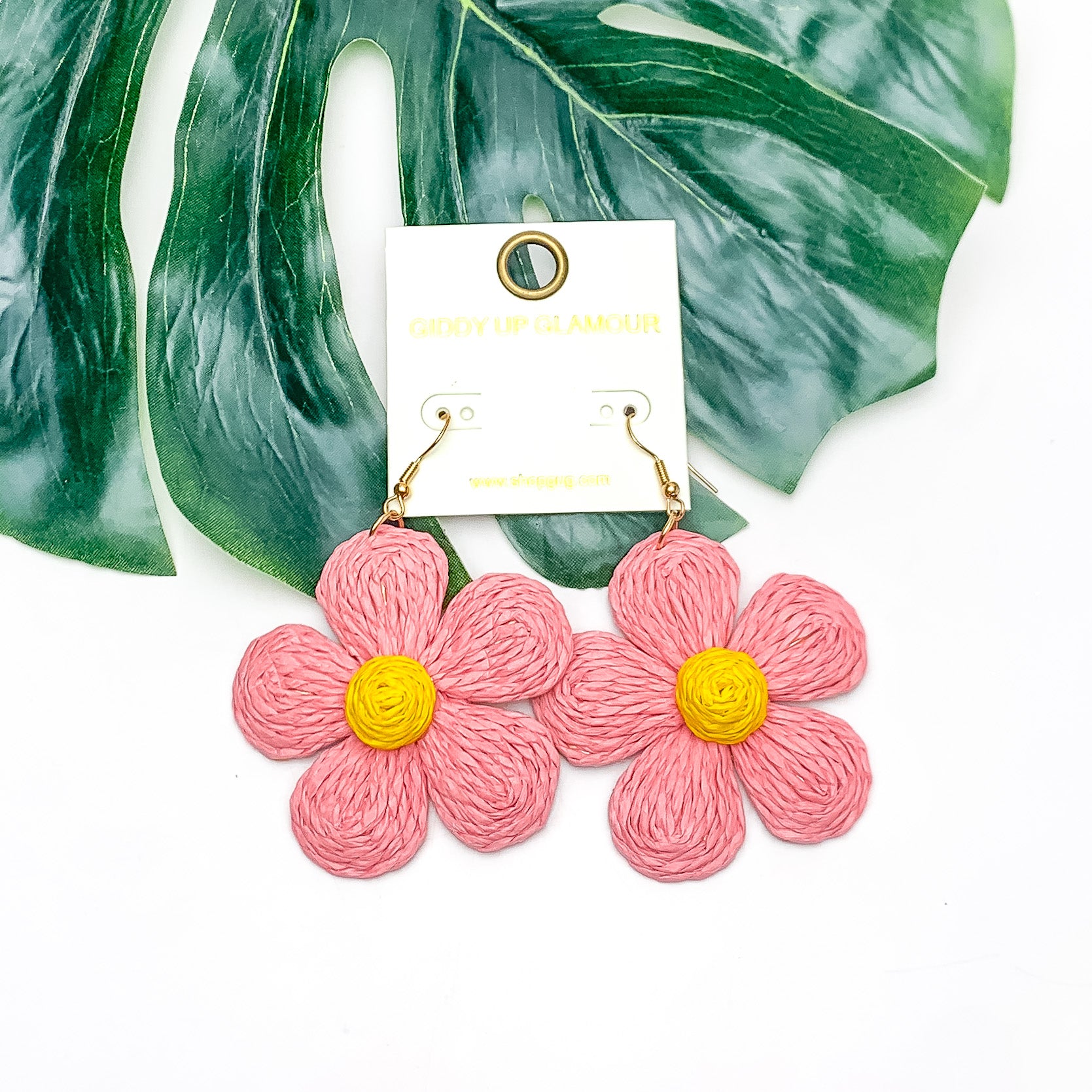 Darling Daisy Raffia Wrapped Flower Earrings in Purple. Pictured on a white background with the earrings laying on a large leaf.