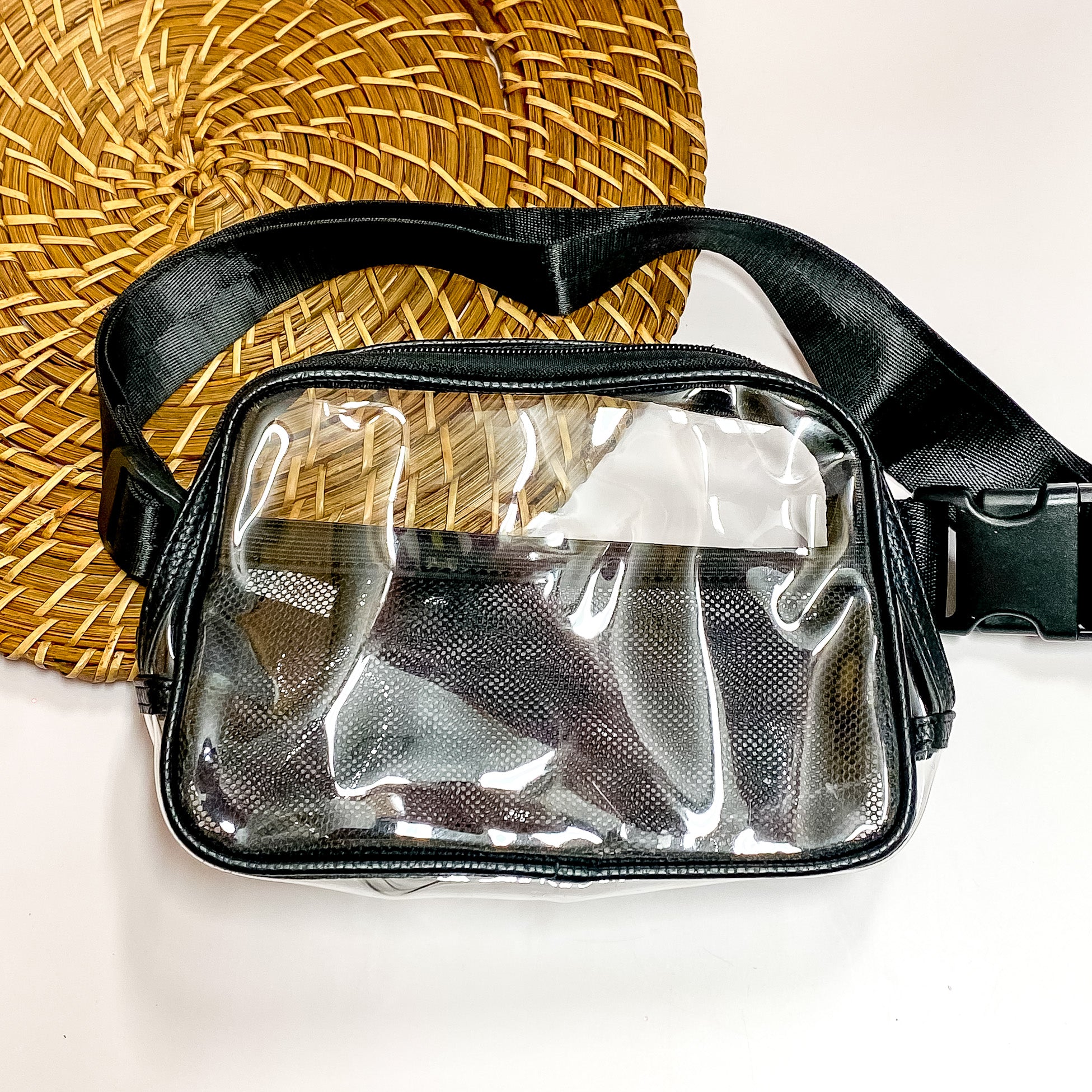 Pictured is a rectangle clear fanny pack with a black outline. This bag also includes a black strap, black accents, and black mesh pockets. This bag is pictured on a white and brown patterned background. 