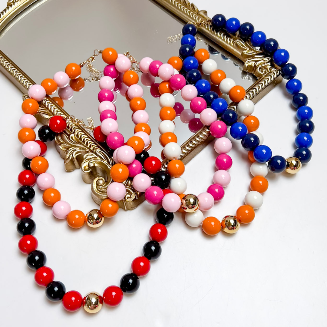 Several bold beaded necklaces in red, black, orange, pink, white and cobalt blue with gold accent beads, layered on an ornate gold mirror frame.