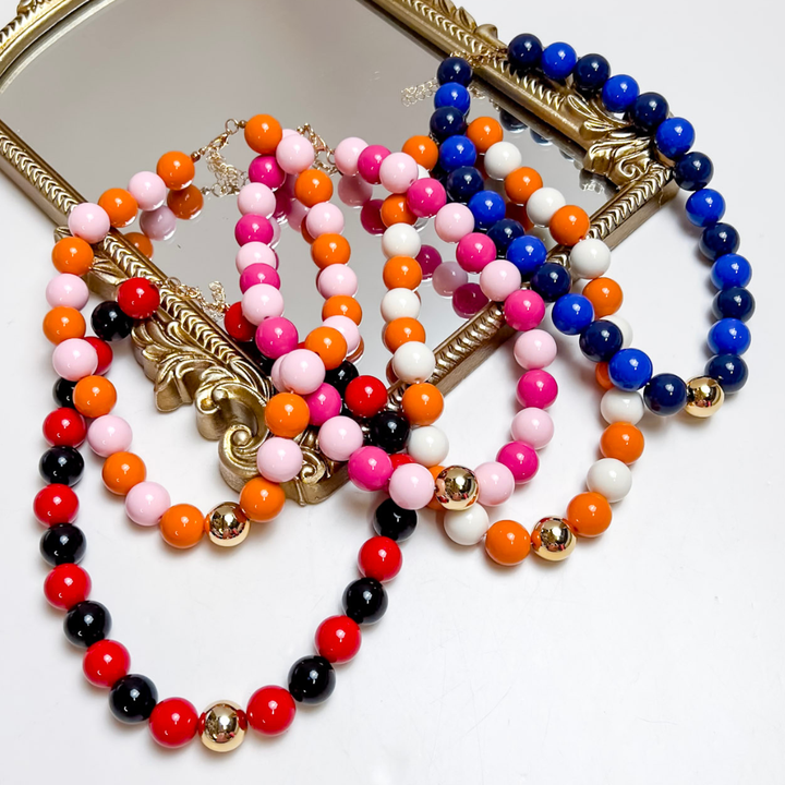 Several bold beaded necklaces in red, black, orange, pink, white and cobalt blue with gold accent beads, layered on an ornate gold mirror frame.