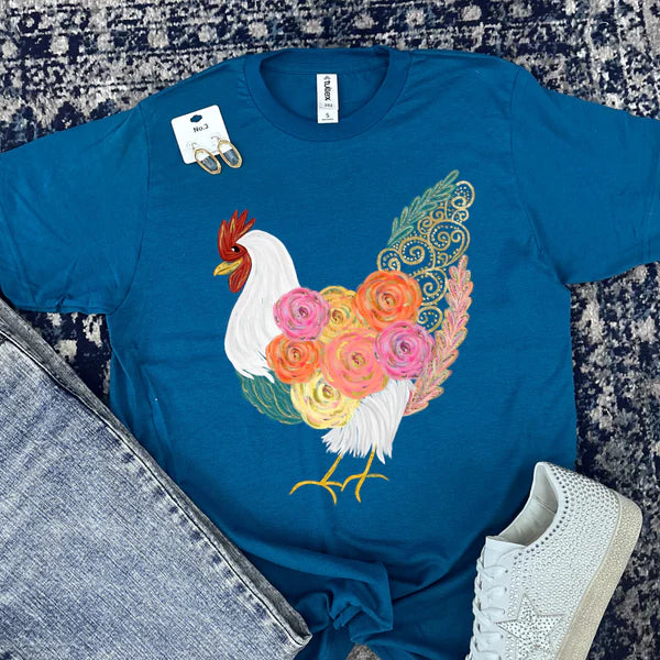 Blue t-shirt with a colorful rooster design on a textured surface with jeans and sneakers.