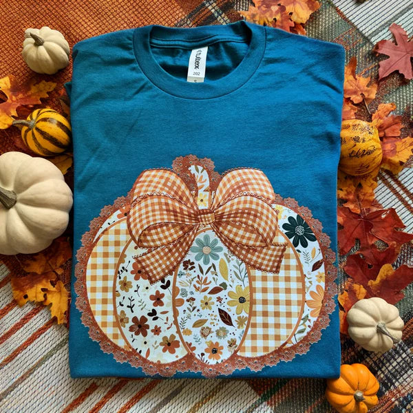 Teal t-shirt with a decorative pumpkin design on a fall-themed background with pumpkins and leaves.