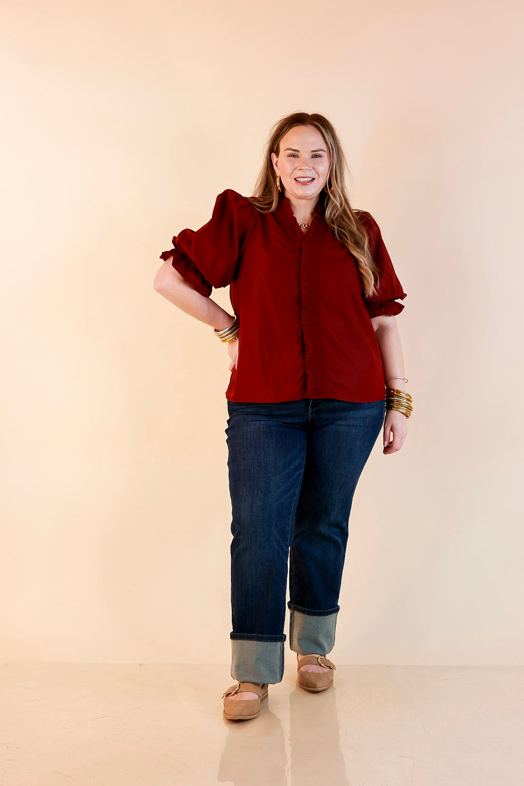 Model wearing a maroon button up top with smocked ruffle trim along the neckline and placket, puff sleeves, cuffed dark wash jeans and tan buckle flats.