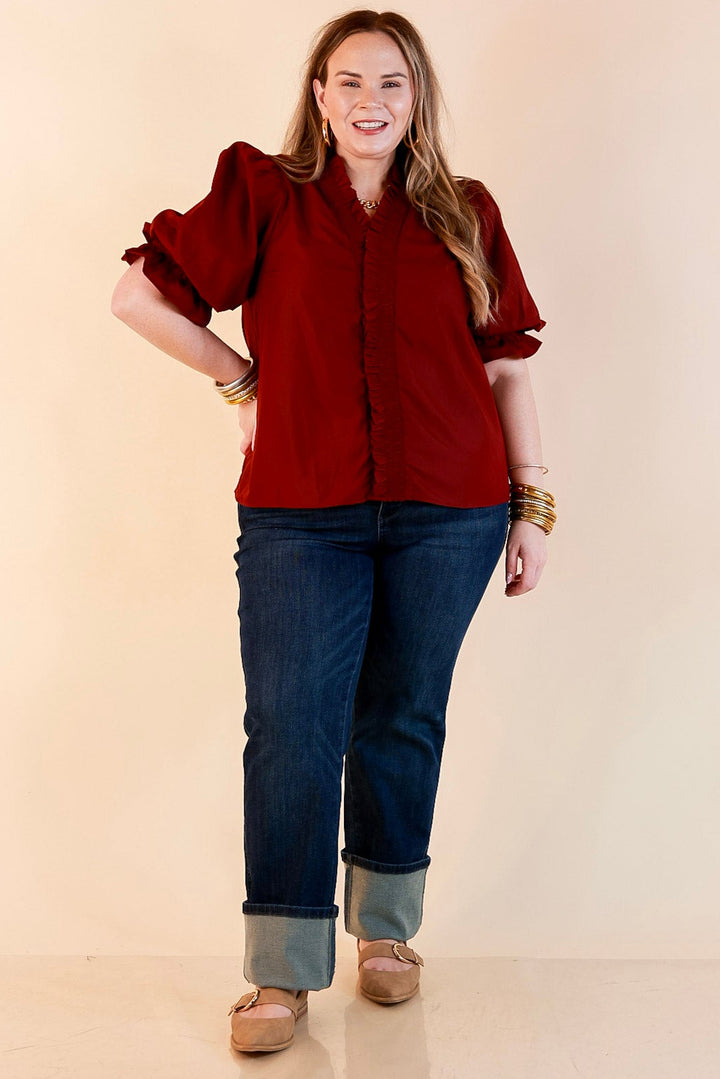 Model wearing a maroon button up top with smocked ruffle trim along the neckline and placket, puff sleeves, cuffed dark wash jeans and tan buckle flats.