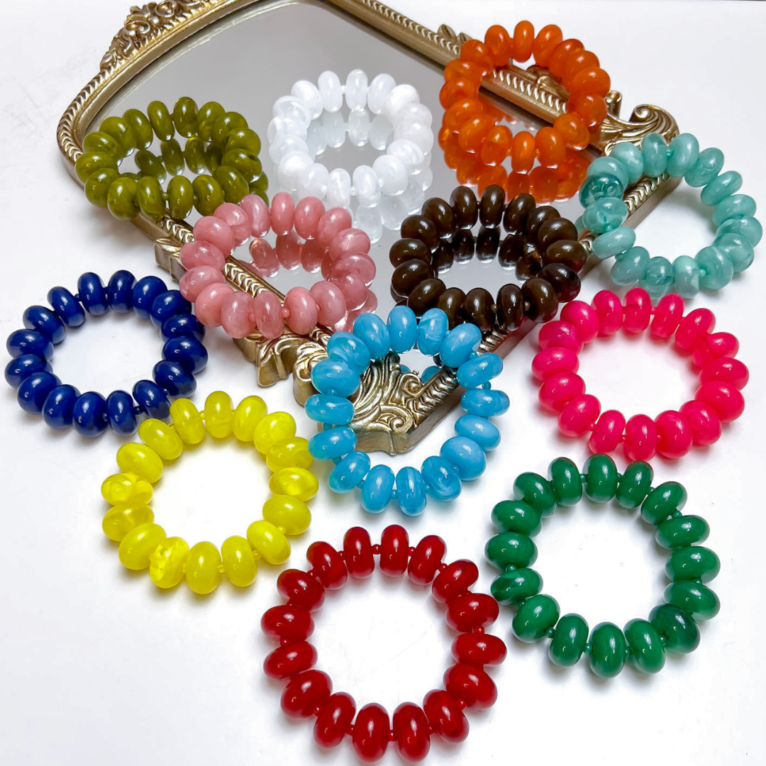 A collection of colorful beaded spiral stretch bracelets arranged on a white surface with an ornate gold mirror in the background. The bracelets come in a variety of colors including blue, yellow, red, green, pink, brown, white, orange, and light blue.