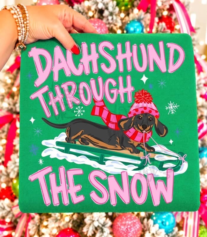 Girly Dachshund Through the Snow Christmas graphic tee or sweatshirt on kelly green with a dachshund in a pink beanie sledding under pink Dachshund Through the Snow lettering