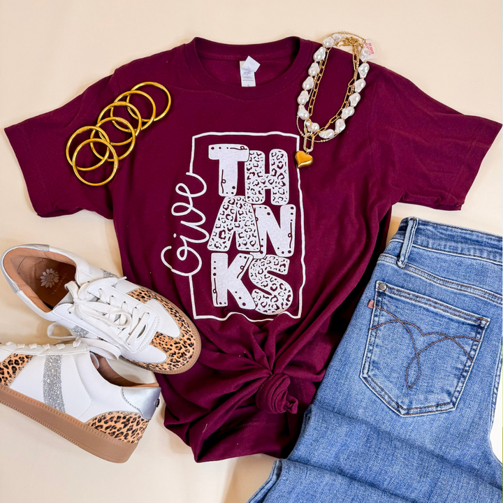 Maroon t-shirt with 'Give Thanks' design, white sneakers, gold bracelets, and blue jeans on a beige background
