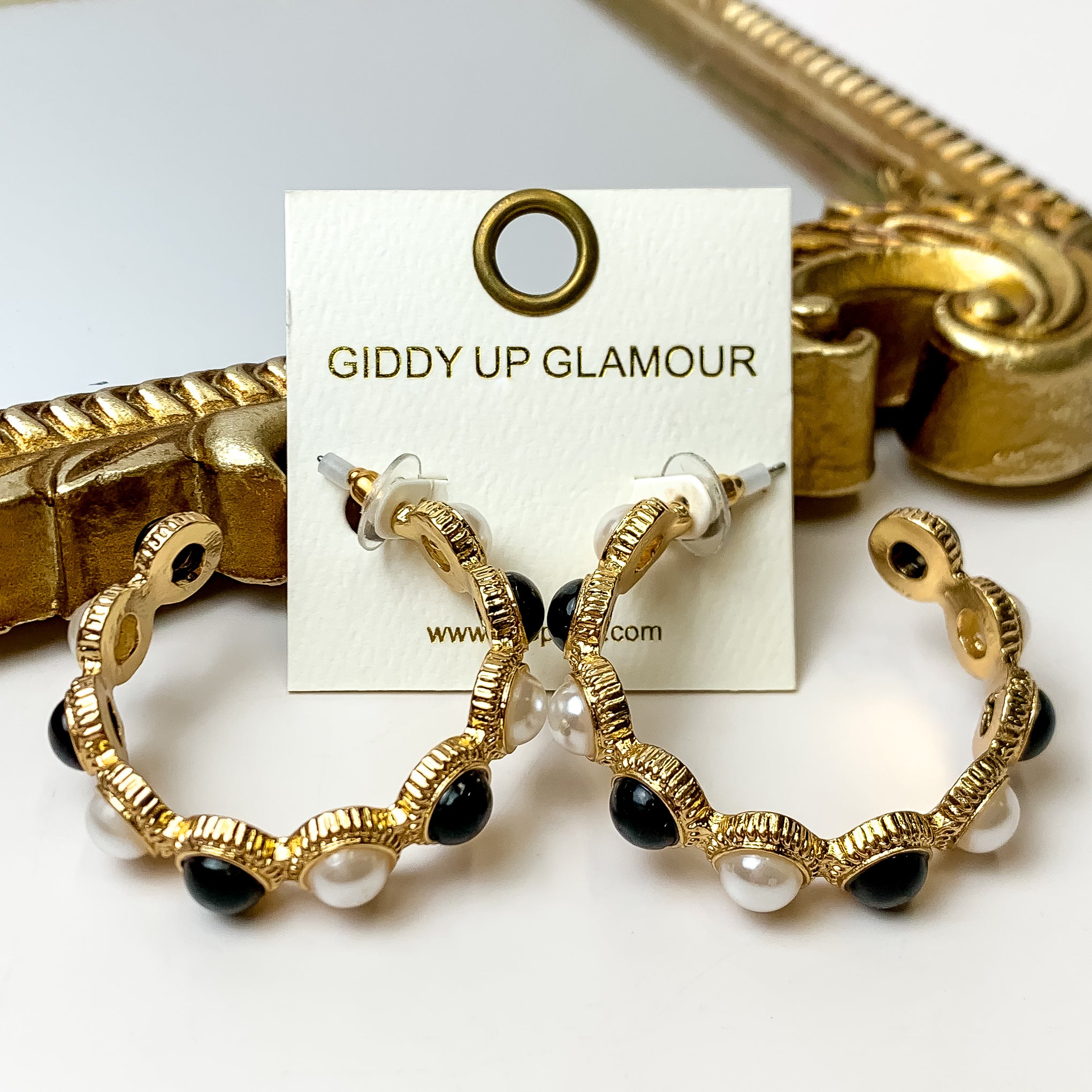 These Gold Tone Pearl Beaded Hoop Earrings in Black and Ivory are placed on an ivory  earring card. They are pictured on a white background with a gold mirror.
