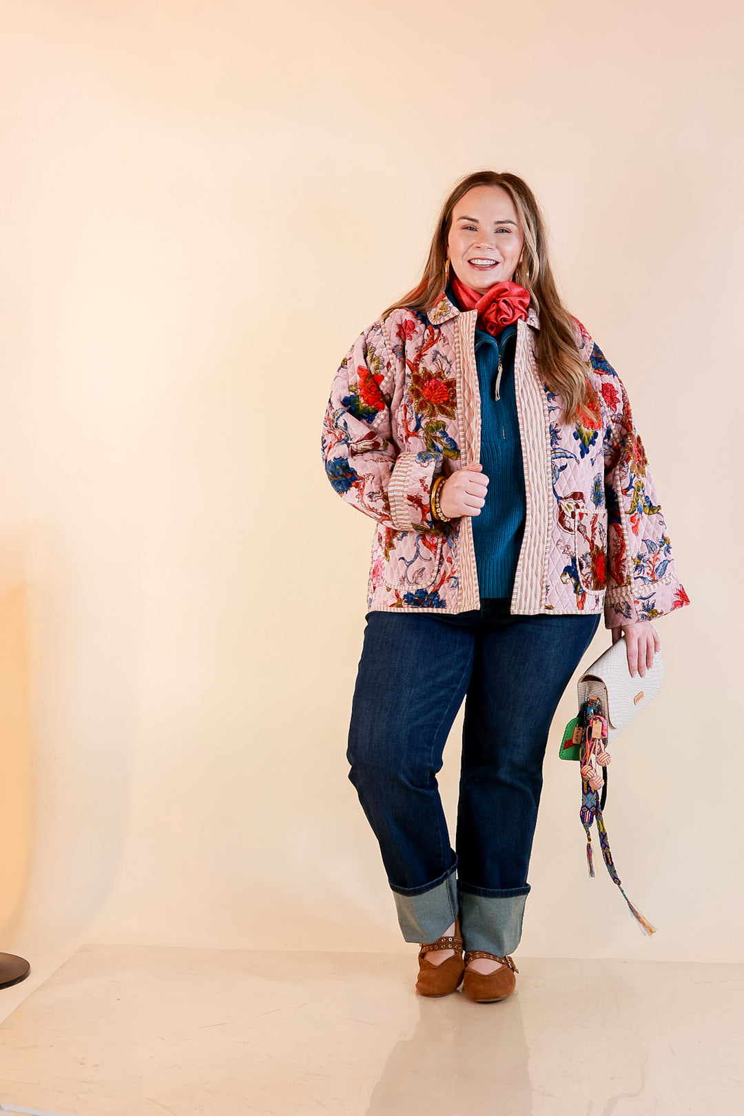 Model wearing a blush pink quilted velvet floral jacket over a teal half-zip sweater with a coral scarf, dark cuffed jeans and brown flats.
