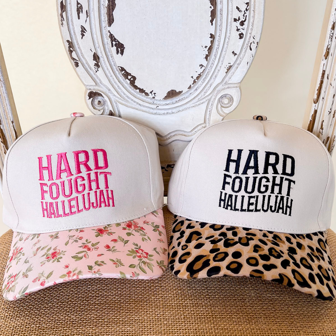Two hats with 'Hard Fought Hallelujah' text, one floral and one leopard print, on a textured surface.