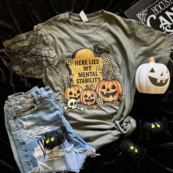 Green t-shirt with Halloween design featuring pumpkins and text, paired with denim shorts and decorative items on a dark background.