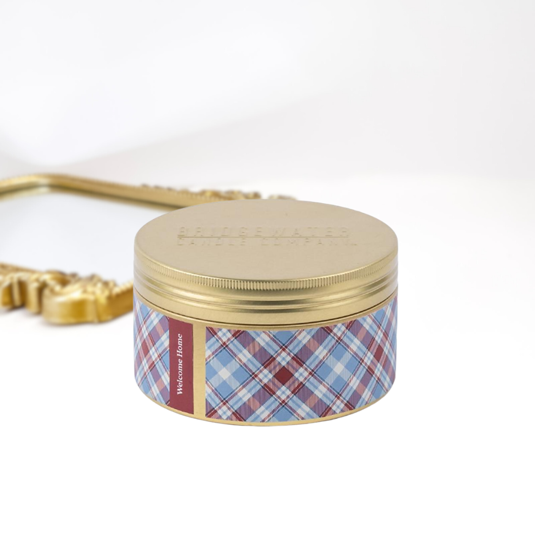A solid white background with a gold mirror and a 3-wick gold tin candle. A maroon and blue plaid print around the tin.