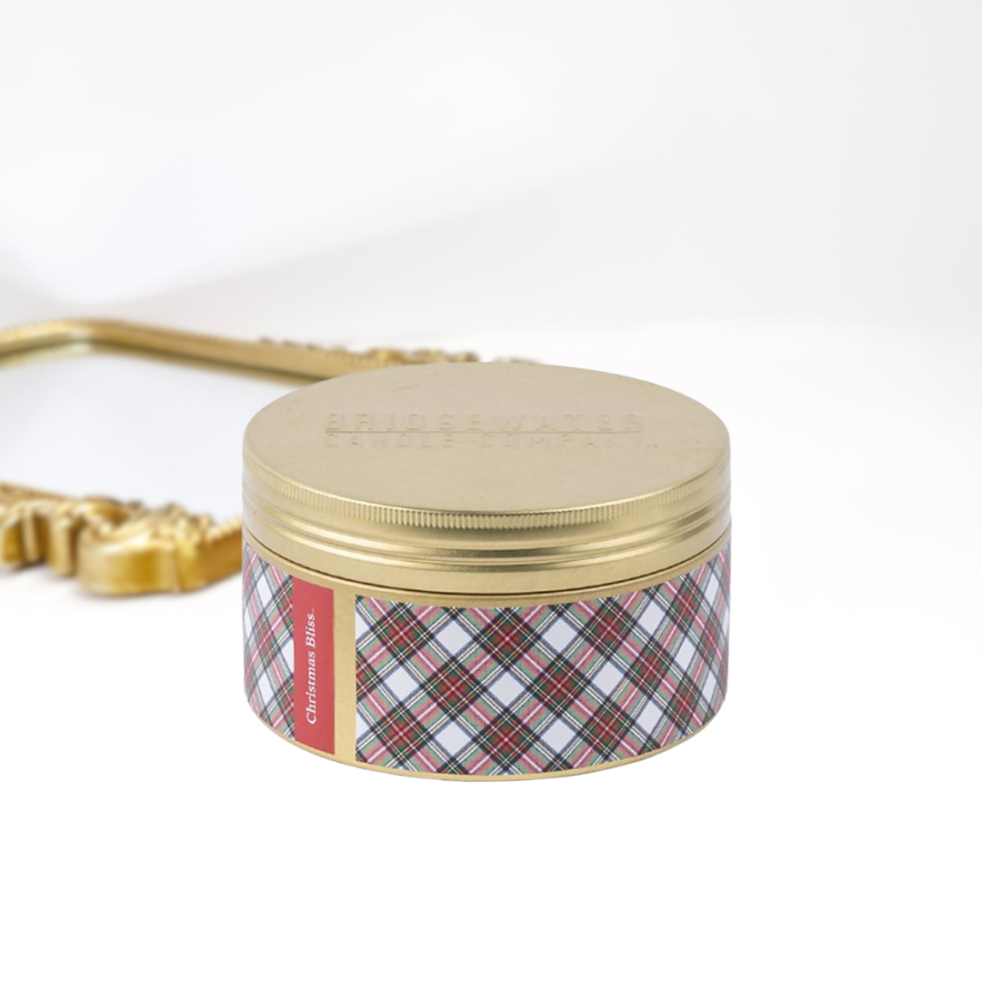 A solid white background with a gold mirror and a 3-wick gold tin candle. A green, red, and white plaid print around the tin.
