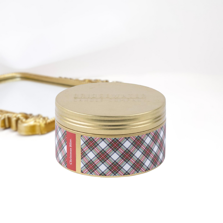 A solid white background with a gold mirror and a 3-wick gold tin candle. A green, red, and white plaid print around the tin.
