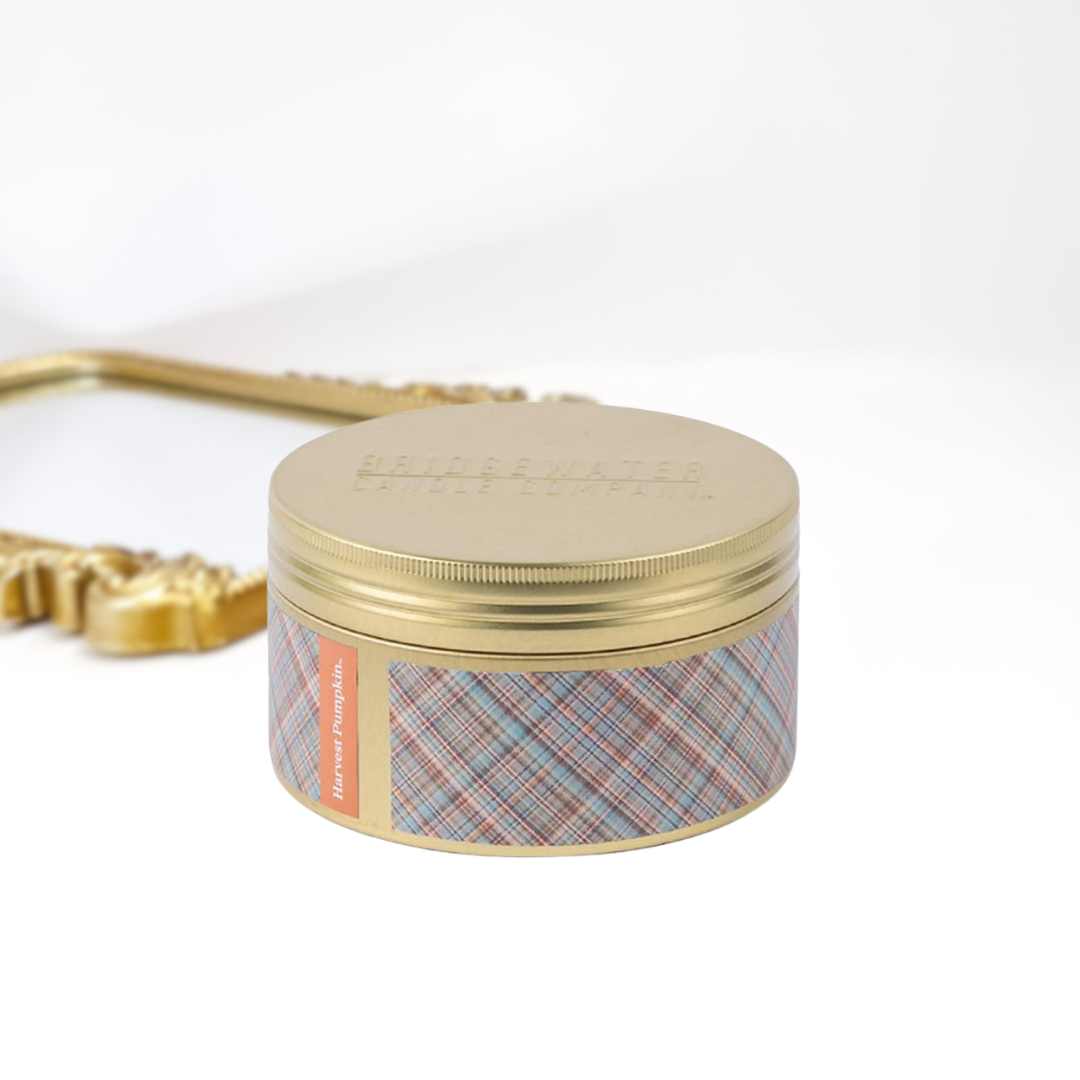 A solid white background with a gold mirror and a 3-wick gold tin candle. A orange and blue plaid print around the tin.