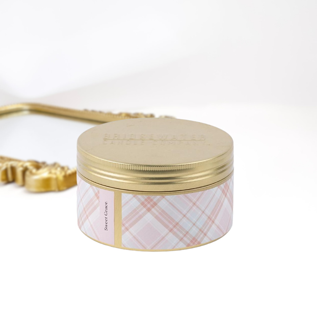 A solid white background with a gold mirror and a 3-wick gold tin candle. A pink and white plaid print around the tin.
