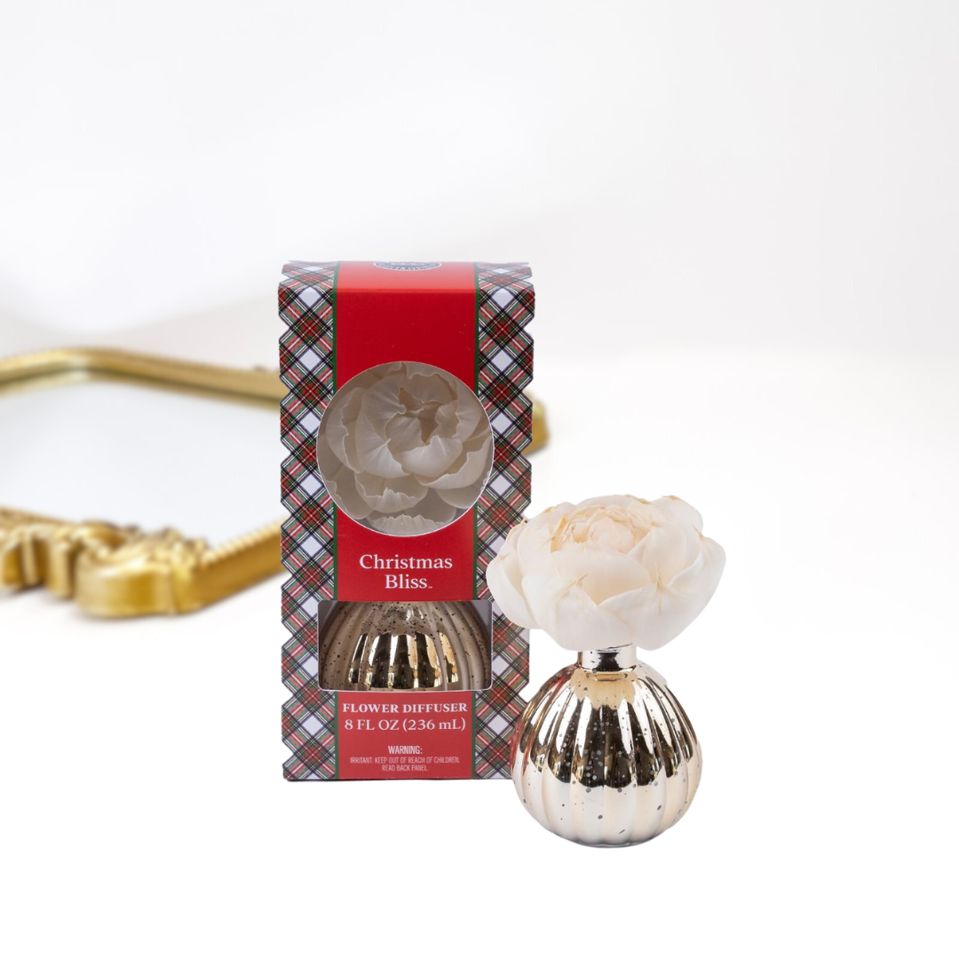 A solid white background with a gold mirror. Upfront is a packaged and unpackaged flower diffuser in the scent christmas bliss by bridgewater candle company.