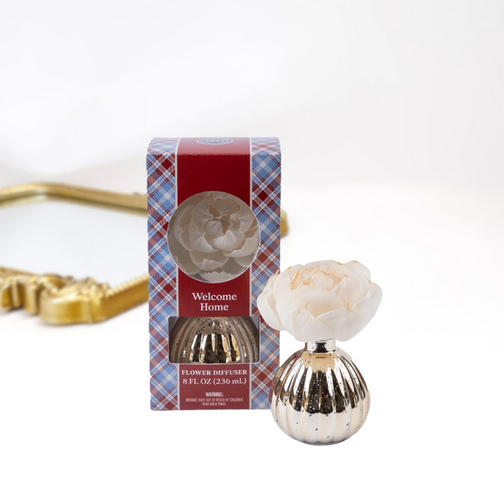 A solid white background with a gold mirror. Upfront is a packaged and unpackaged flower diffuser in the scent welcome home by bridgewater candle company.